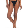 A person stands with one leg slightly crossed in front of the other, modeling the Sanctuary Clothing Double Trouble V Hipster Bikini Bottom in black. The skin tone is medium-dark, and the individual's torso is not visible. The background is white, and the person is barefoot.