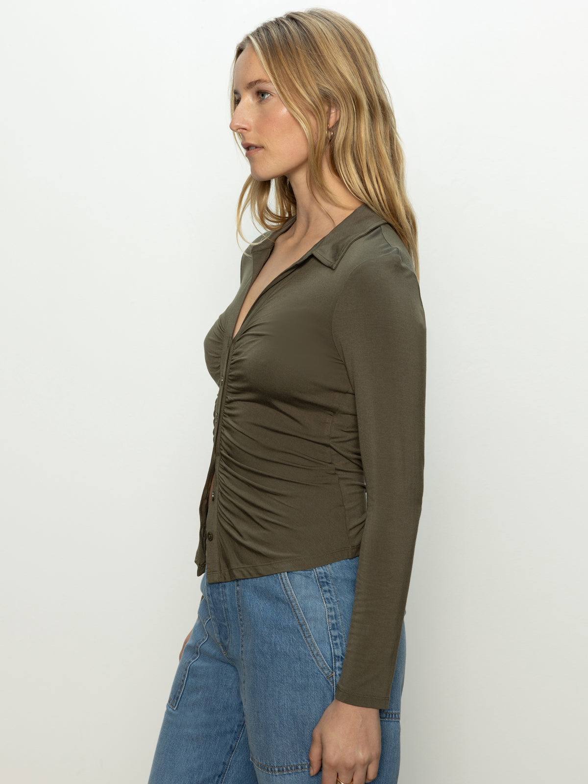 A person with long blonde hair stands in profile against a plain background, wearing Sanctuary Clothing's Dreamgirl Button Up Top in Kalamata and light blue jeans.