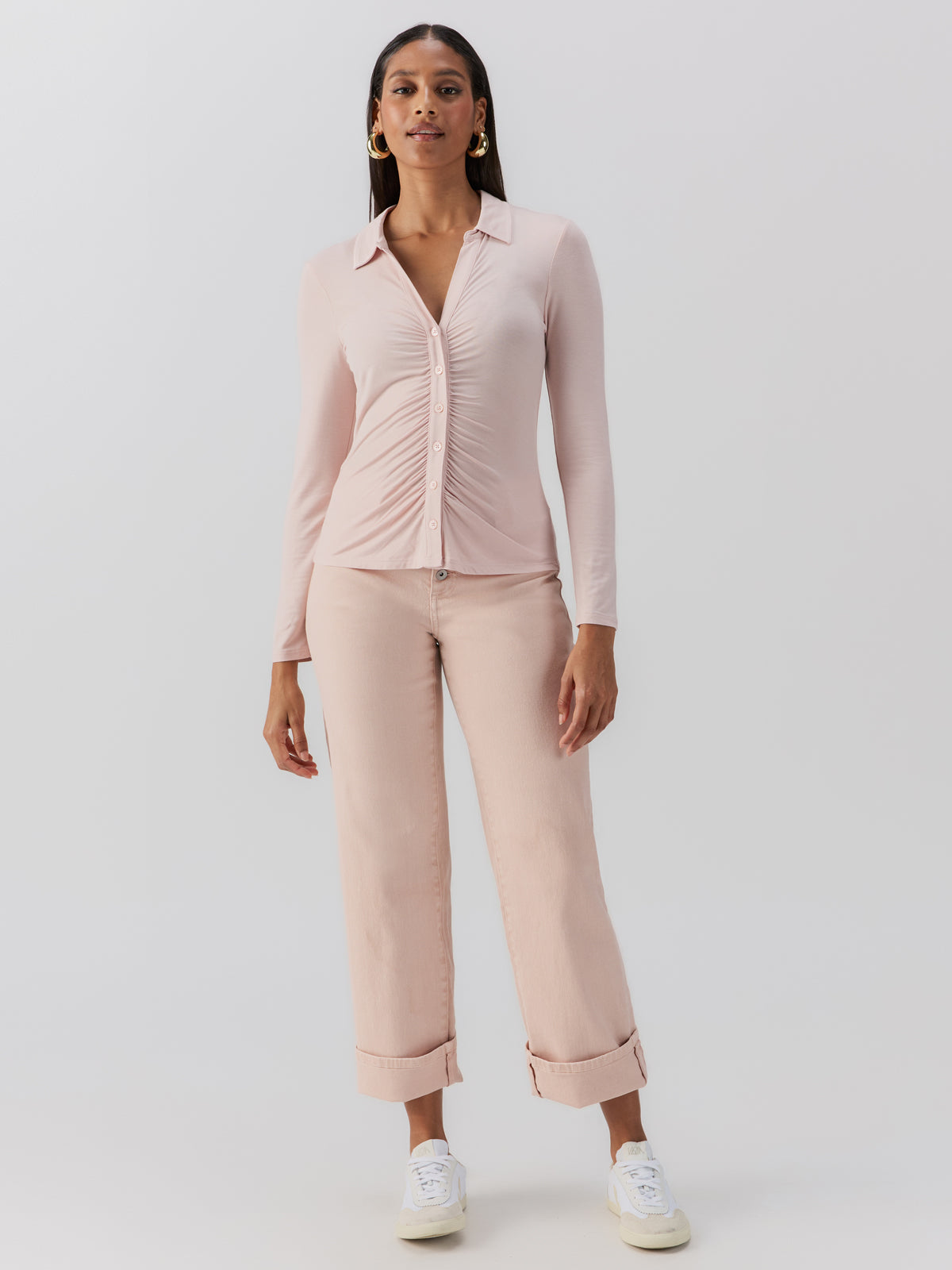 A woman is posed against a plain background, dressed in the "dreamgirl button up light rosewood" by Sanctuary Clothing. She wears matching light pink pants, white sneakers, and large gold hoop earrings. Her hair is long and straight.