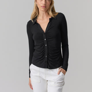 A person with long blonde hair is standing against a plain background. They are wearing the "Dreamgirl Button Up Black" by Sanctuary Clothing paired with white pants. One hand is in their pocket, and they have a neutral expression.