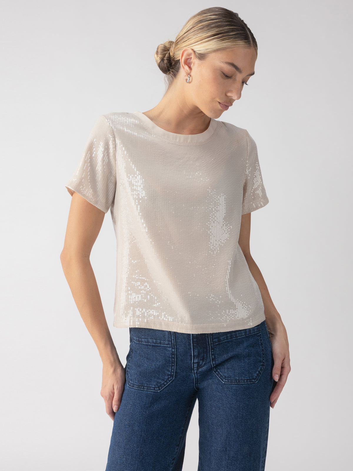 A person with styled hair poses in the Perfect Sequin Tee Frosted Almond by Sanctuary Clothing and blue jeans. With one hand resting on their hip, they look downwards against a plain, light gray background.