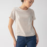 A person with styled hair poses in the Perfect Sequin Tee Frosted Almond by Sanctuary Clothing and blue jeans. With one hand resting on their hip, they look downwards against a plain, light gray background.