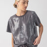 A person dressed in the Sanctuary Clothing Perfect Sequin Tee Gunmetal, which sparkles in various shades of gray and has short sleeves, is paired with matching pants. Their hair is styled neatly in a bun, and they are posing slightly to the side against a simple, light gray background.