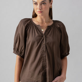 A person with long brown hair and subtle makeup is wearing the Sanctuary Clothing Bubble Sleeve Boho Blouse in Mud Bath, paired with beige pants. They are standing against a plain light gray background, gazing slightly to the side with a calm expression.