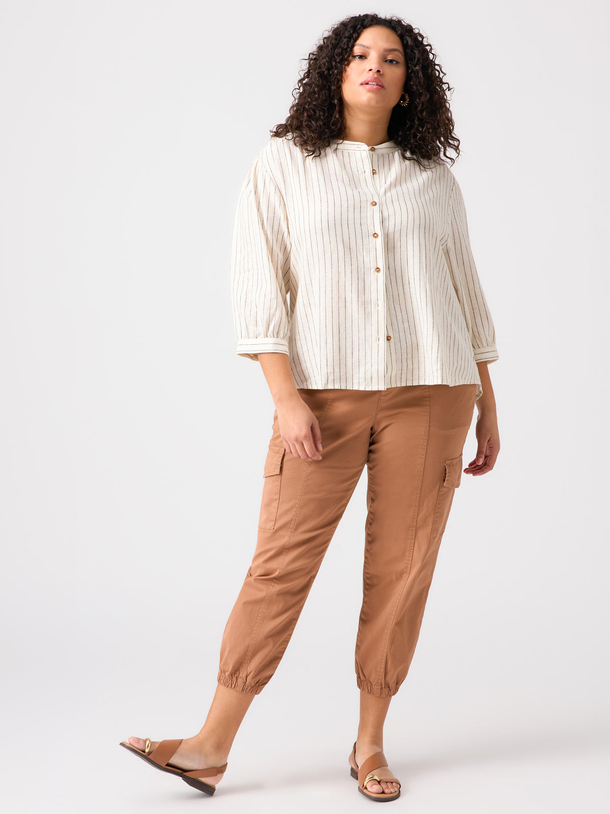 A person with curly hair is wearing The Femme Shirt Birch Stripe from the Inclusive Collection by Sanctuary Clothing, paired with brown cargo pants and white sandals. They are standing against a plain white background.