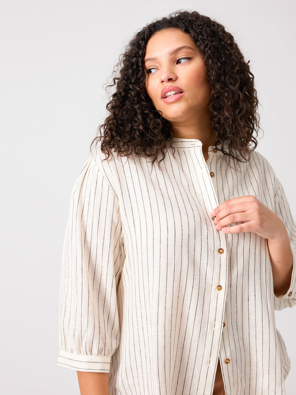 A person with curly hair is wearing The Femme Shirt Birch Stripe from Sanctuary Clothing's Inclusive Collection. They are looking to the side with a neutral expression, with one hand raised towards the shirt's buttons. The background is plain and light-colored.