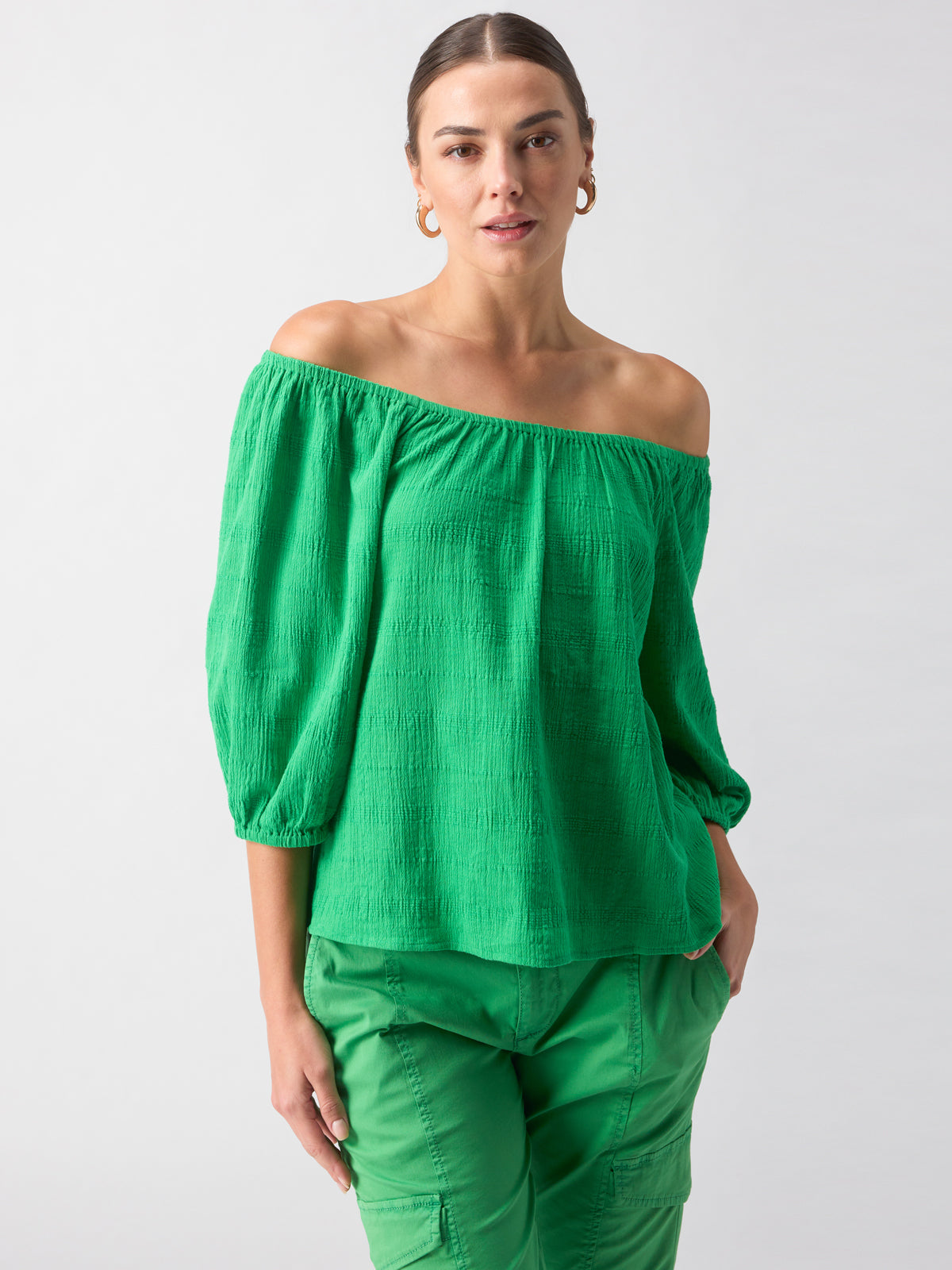 A woman stands confidently against a plain background, wearing the Sanctuary Clothing Beach To Bar Blouse Green Goddess with three-quarter sleeves and matching green cargo pants. She has her hair pulled back and is accessorized with gold hoop earrings.