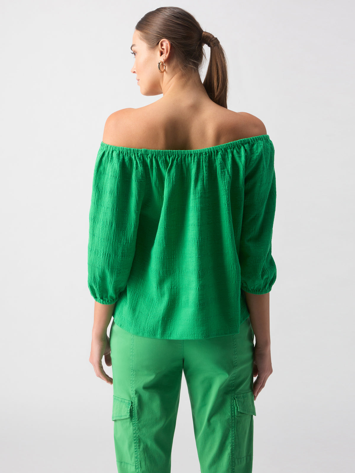 A woman stands facing away, wearing the Beach To Bar Blouse Green Goddess by Sanctuary Clothing, an off-the-shoulder top with three-quarter sleeves. She pairs it with green cargo pants. Her hair is tied back in a ponytail, and she adorns hoop earrings. The background is plain and light-colored.