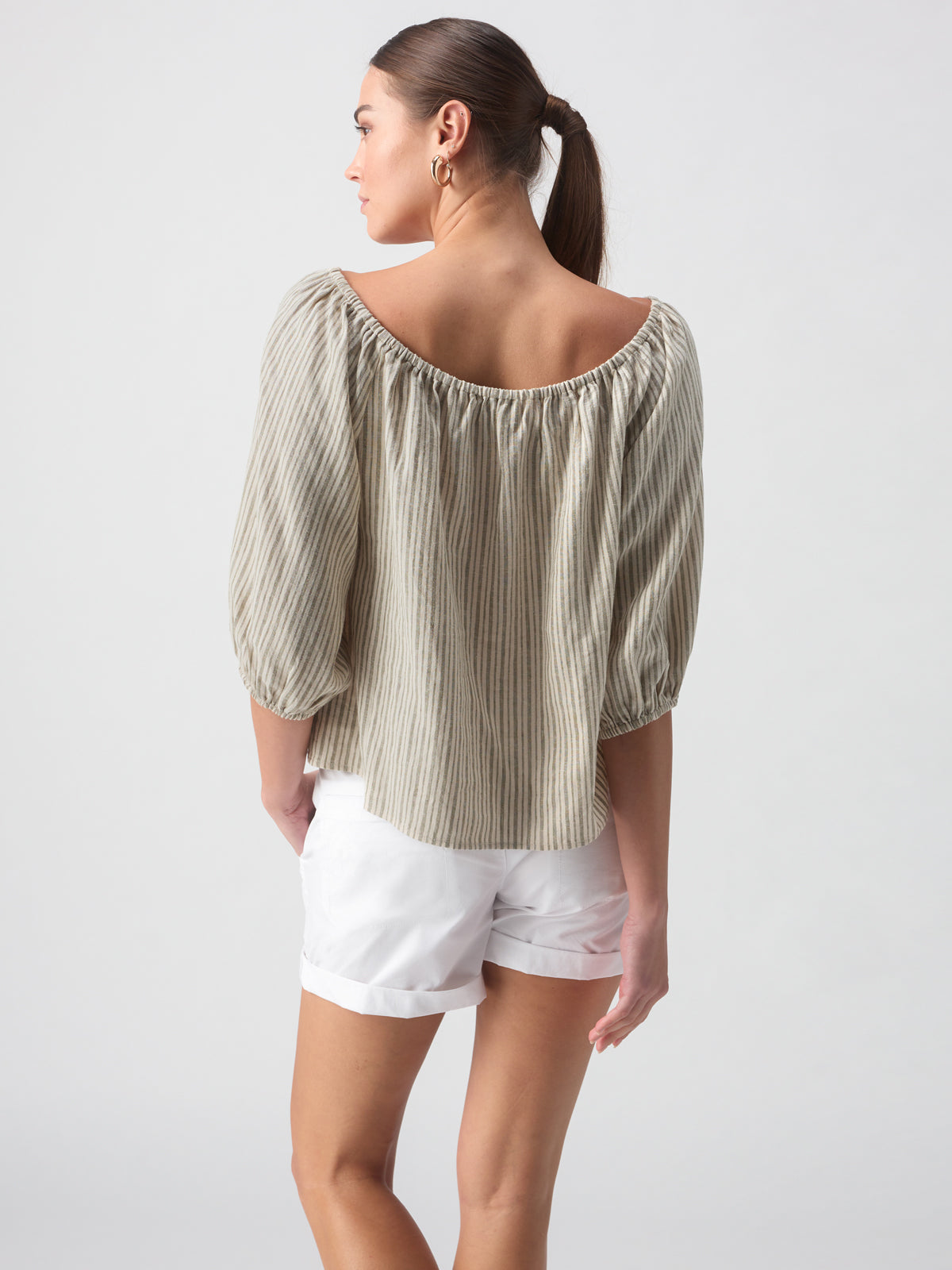 A woman with long hair in a ponytail is wearing the Sanctuary Clothing Beach To Bar Blouse Eco Olive Stripe, paired with white shorts. She is standing with her back to the camera, showcasing the details of her outfit against a plain background.