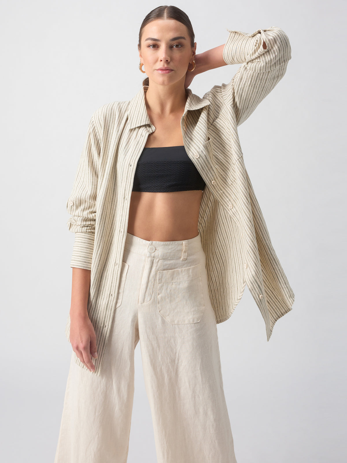 Person with long hair tied back, wearing the Sanctuary Clothing Long Line Pocket Shirt Eco Olive Stripe over a black crop top and high-waisted, wide-leg beige pants, standing against a plain grey background. She has one hand resting on her hip and the other behind her head.