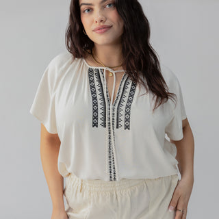 A person wearing the Golden Dream Top Birch from Sanctuary Clothing's Inclusive Collection, featuring black embroidered patterns along the neckline and front, paired with light-colored shorts. They are standing in front of a neutral background, with their hair down and a gentle expression on their face.