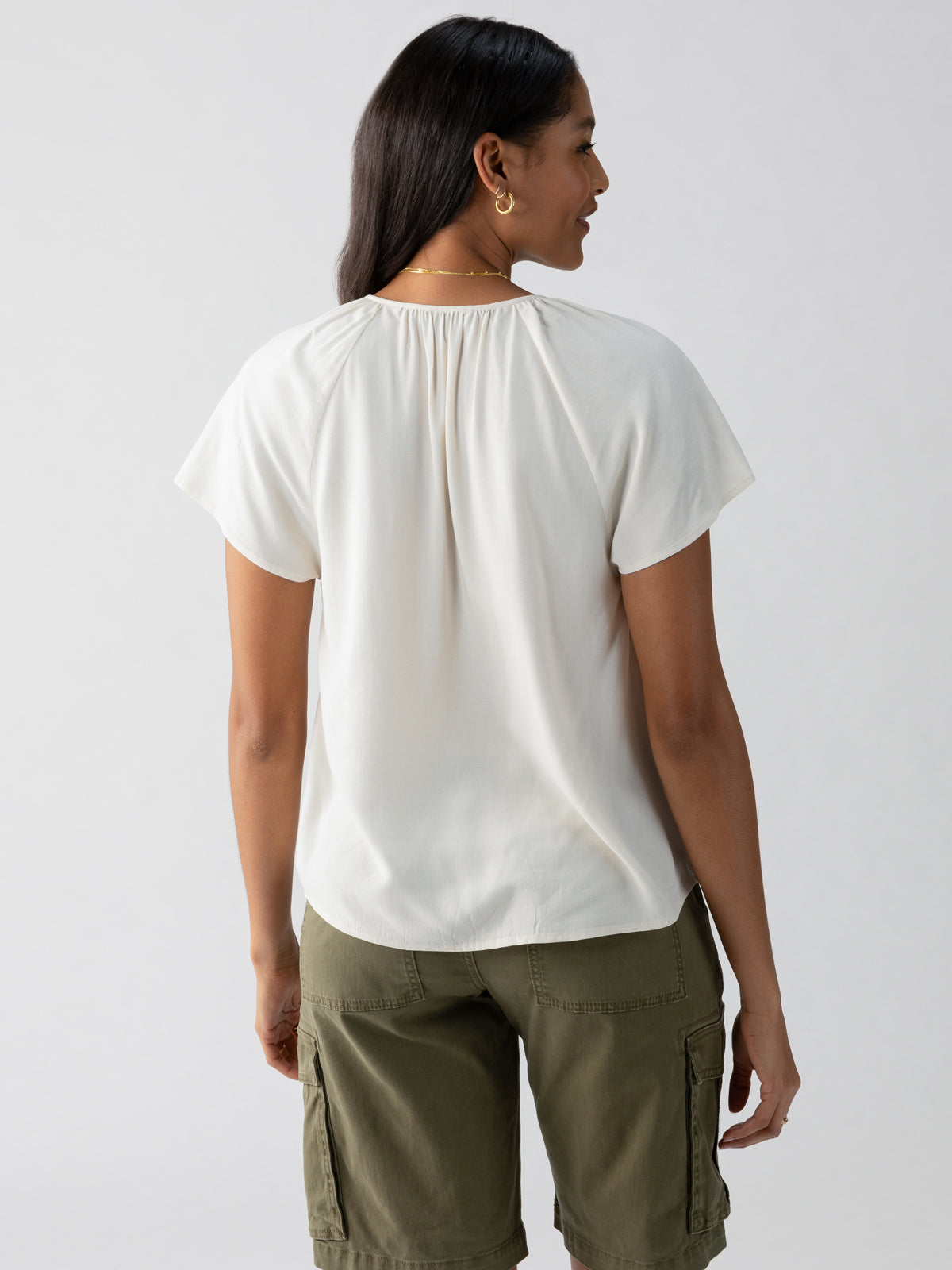 A person with long dark hair is shown from the back, wearing a Sanctuary Clothing "Golden Dream Top Birch" short-sleeved white blouse and olive green cargo shorts. The background is plain and light-colored.