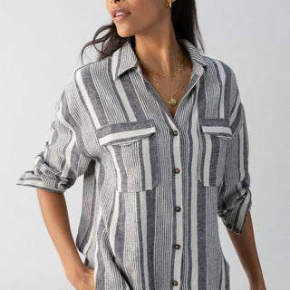 A person wearing the Sanctuary Clothing Pocket Shirt Varigated Stripe, a long-sleeved, button-down shirt with vertical gray and white stripes. They have their hands in their pockets and are looking to the side. They have long, dark hair and are accessorized with gold necklaces and earrings.