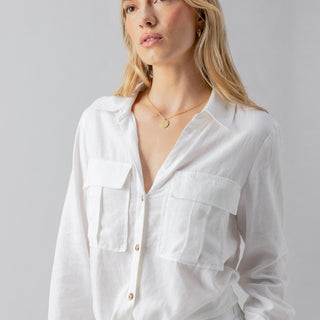 A woman with long blonde hair, adorned in the Utility Pocket Shirt White by Sanctuary Clothing, accessorized with a gold necklace, stands against a plain light gray background. Her calm expression and slight sideways glance complete the serene imagery.