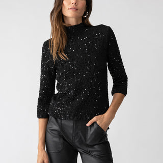 A woman standing against a plain background, wearing the "Almost Holiday Black" long-sleeved top with a sparkly texture and black leather pants by Sanctuary Clothing. Her left hand is in her pocket, and she has long brown hair styled with gentle waves.