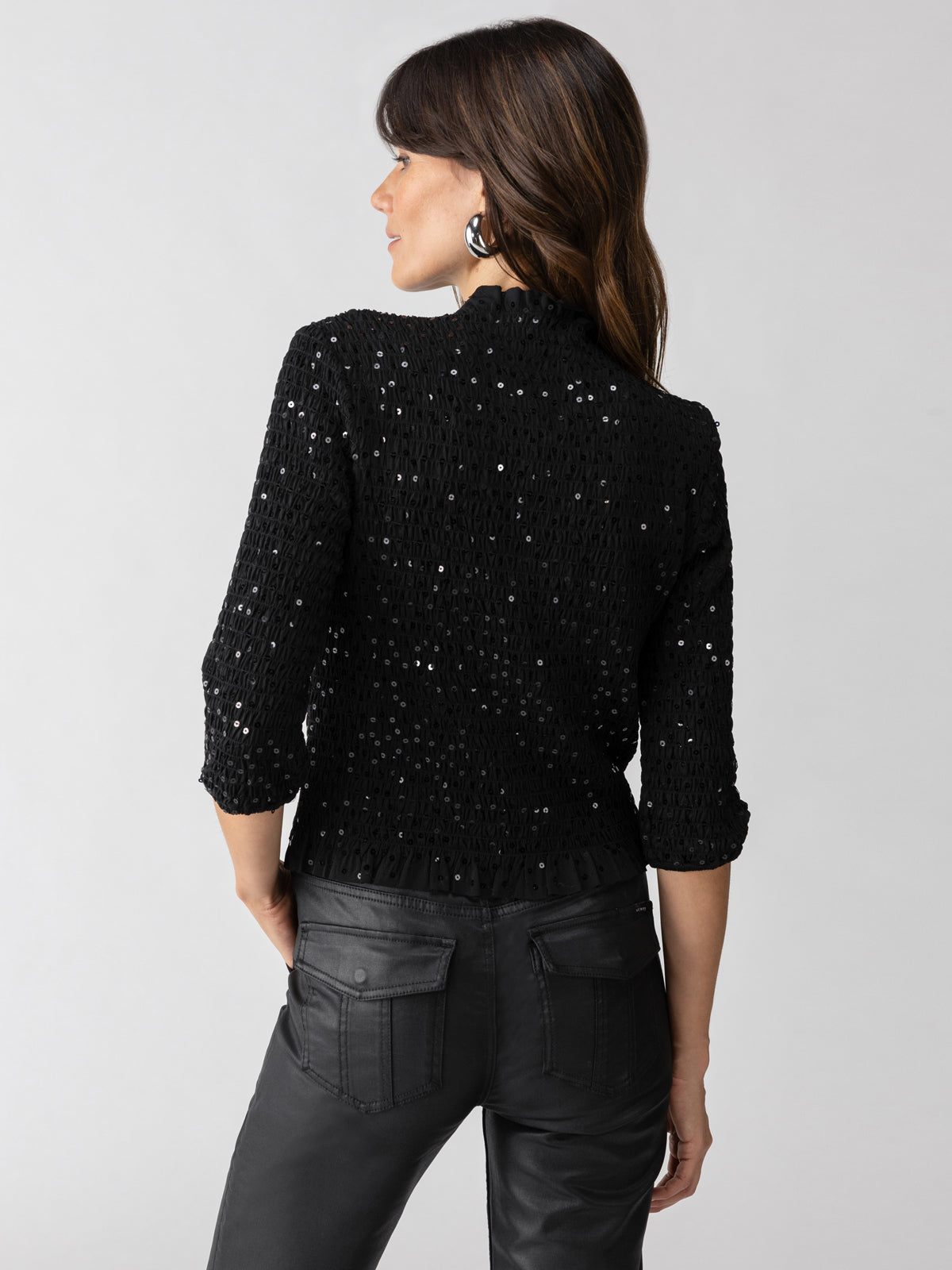 A woman with long brown hair faces away from the camera, wearing the Sanctuary Clothing's Almost Holiday Black textured long-sleeve top and black pants. The top features a subtle sparkle and a high collar as she stands against a plain gray background.