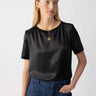 A person with long blond hair is wearing the Timeless Satin Tee Black from Sanctuary Clothing, accompanied by blue jeans and a gold necklace with a pendant. They have one hand in their pocket and are looking away from the camera against a plain, light-colored background.