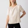 A person with long, light hair stands against a plain background, wearing the Timeless Satin Tee in Toasted Almond by Sanctuary Clothing and black pants. They have a relaxed expression and one hand placed on their hip.