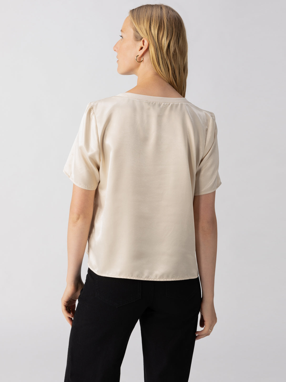 A woman with blonde hair, seen from behind, wearing the Sanctuary Clothing "Timeless Satin Tee" in Toasted Almond paired with black pants. She stands against a plain, light grey background, with her head turned slightly to the side.