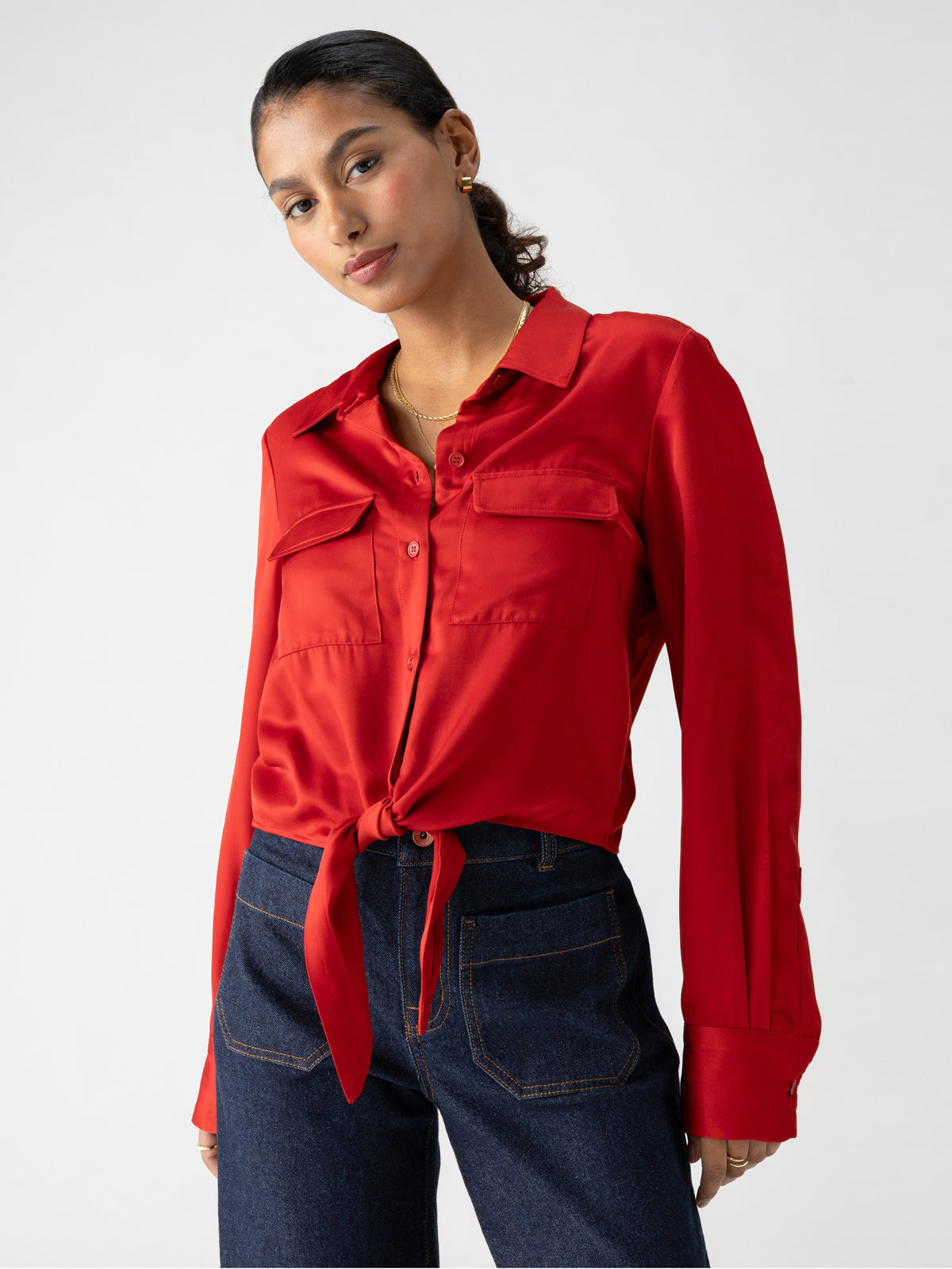 A person with dark hair tied back is wearing a Sanctuary Clothing Lifetime Shirt in Cherry Red, styled with a knot at the waist, paired with dark blue jeans. They have gold hoop earrings and are looking directly at the camera against a plain white background.