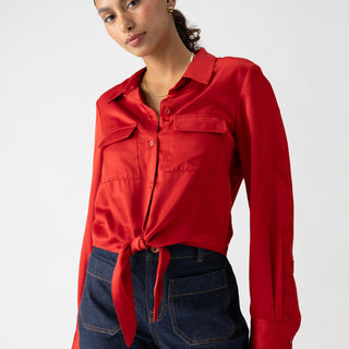 A person with dark hair tied back is wearing a Sanctuary Clothing Lifetime Shirt in Cherry Red, styled with a knot at the waist, paired with dark blue jeans. They have gold hoop earrings and are looking directly at the camera against a plain white background.