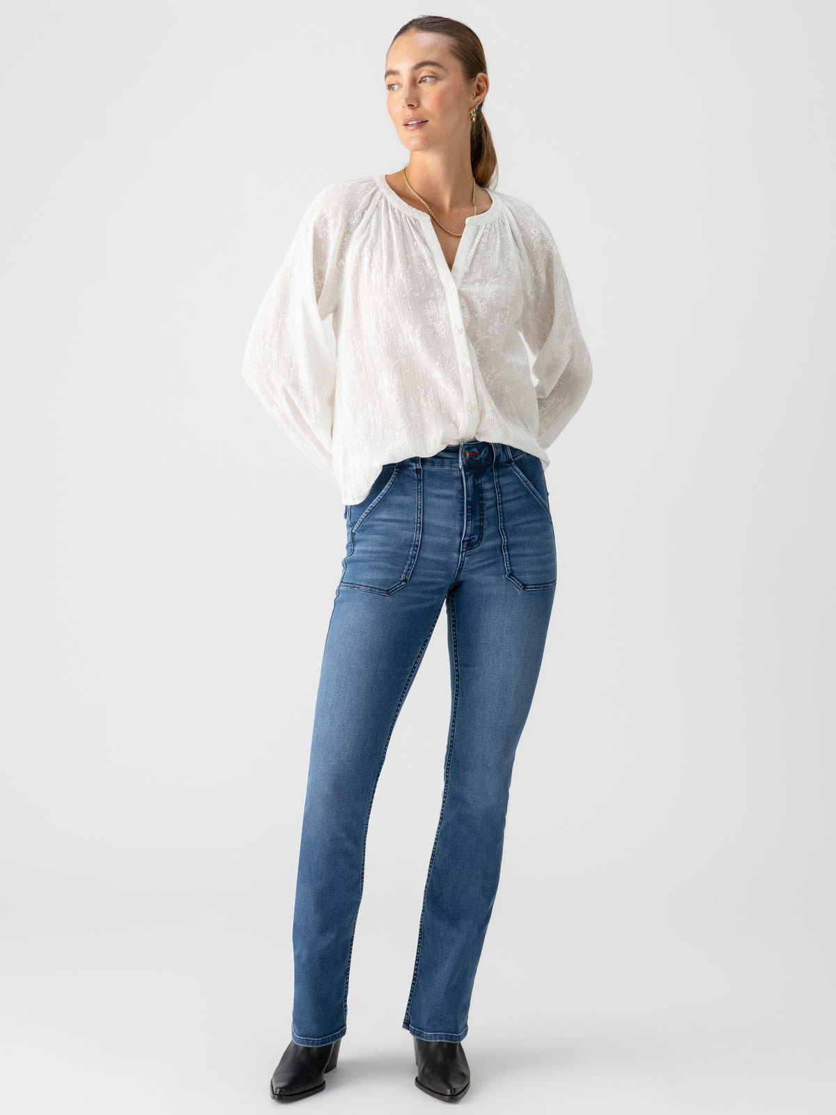 A woman stands against a plain white background, wearing the Shimmer Blouse Chalk from Sanctuary Clothing. The long-sleeved, loose-fitting white blouse is paired with high-waisted blue jeans and black shoes. She looks slightly to her left with her hands in her pockets and her straight hair pulled back.