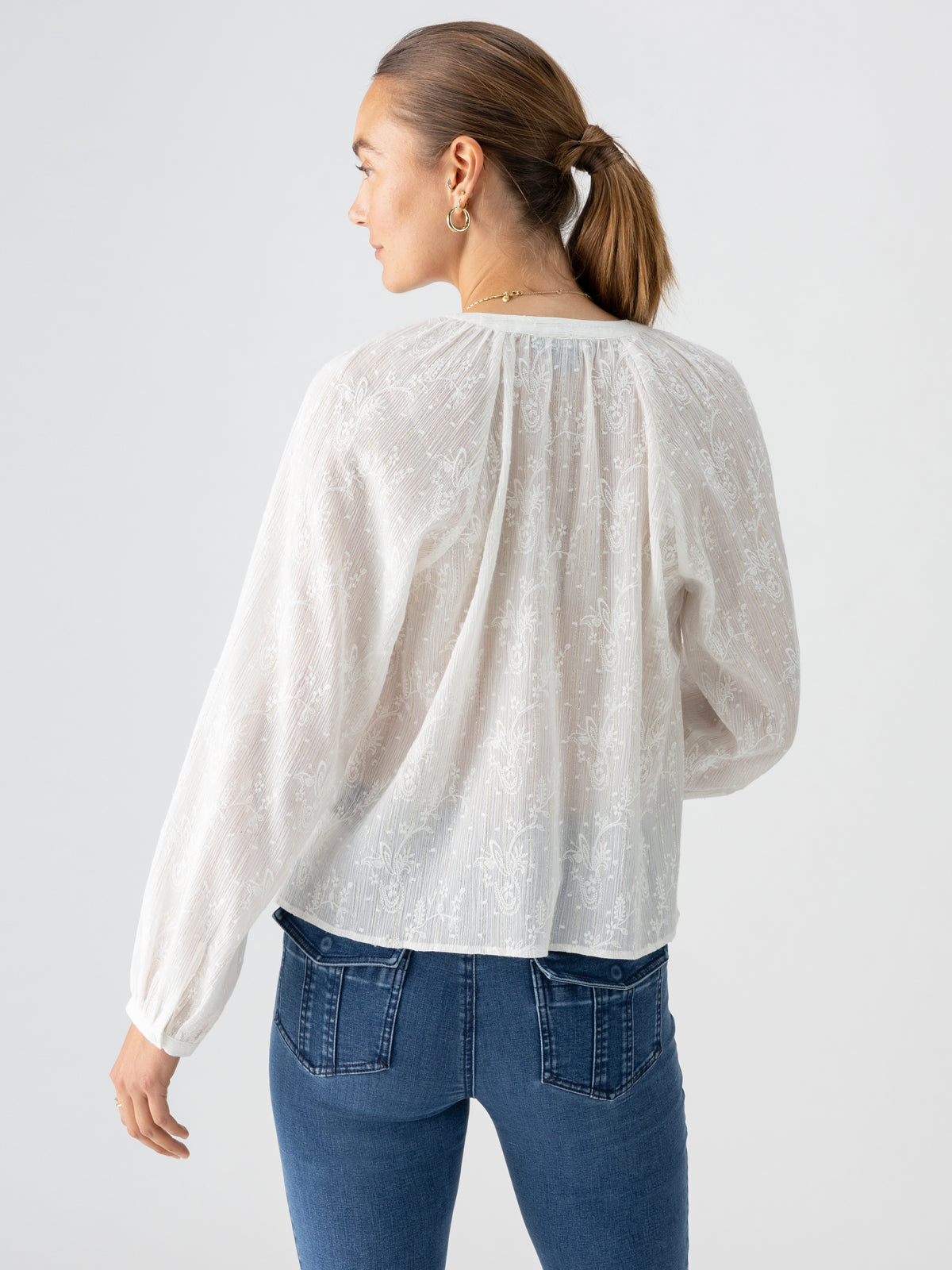 A woman with her hair in a low ponytail is facing away. She wears the Shimmer Blouse Chalk by Sanctuary Clothing, featuring a lightly textured, sheer white design and long sleeves. She pairs it with blue jeans that have detailed stitching on the back pockets. The background is plain and light-colored.