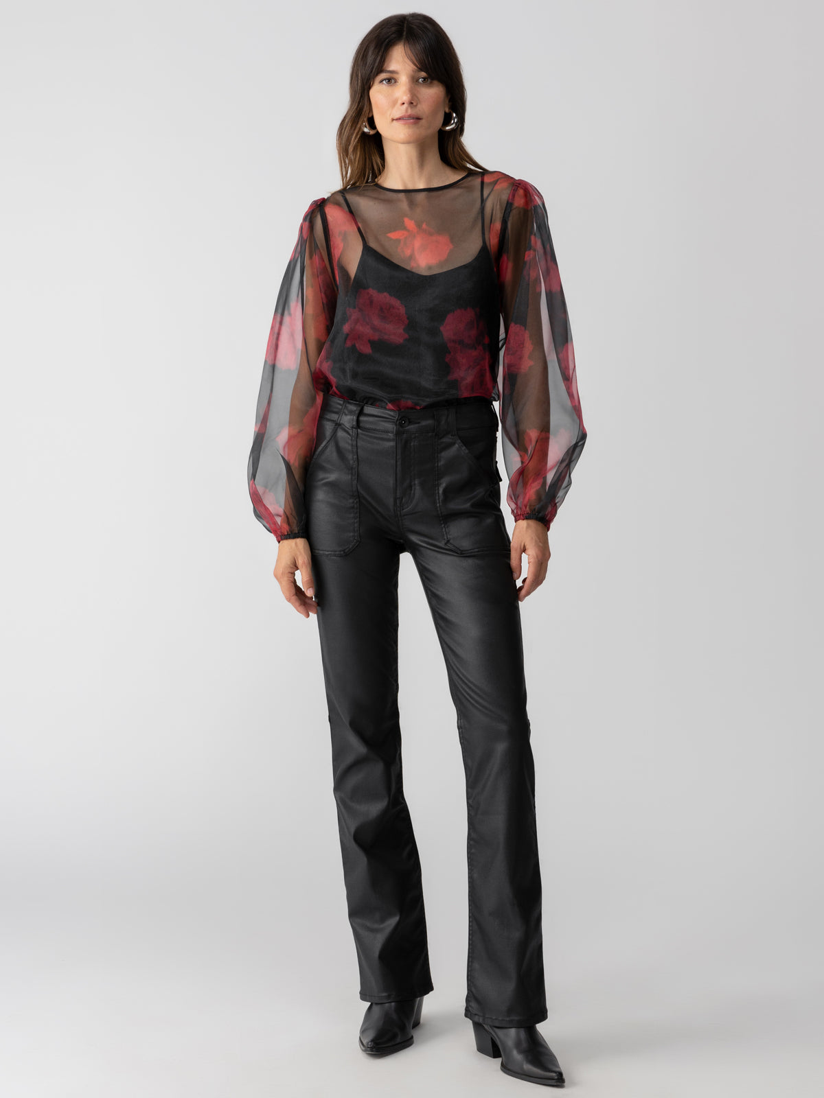 A woman stands confidently, wearing the Moody Sheer Blouse Moody Rose from Sanctuary Clothing, featuring a sheer black design with red floral patterns and long puffy sleeves over a black camisole. She pairs it with black leather pants and black pointed-toe boots. The background is plain and white.