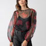 A woman with long brown hair is wearing the Moody Sheer Blouse Moody Rose by Sanctuary Clothing, featuring red floral patterns over a black camisole, paired with black leather pants. She stands against a plain light grey background and accessorizes with large hoop earrings.