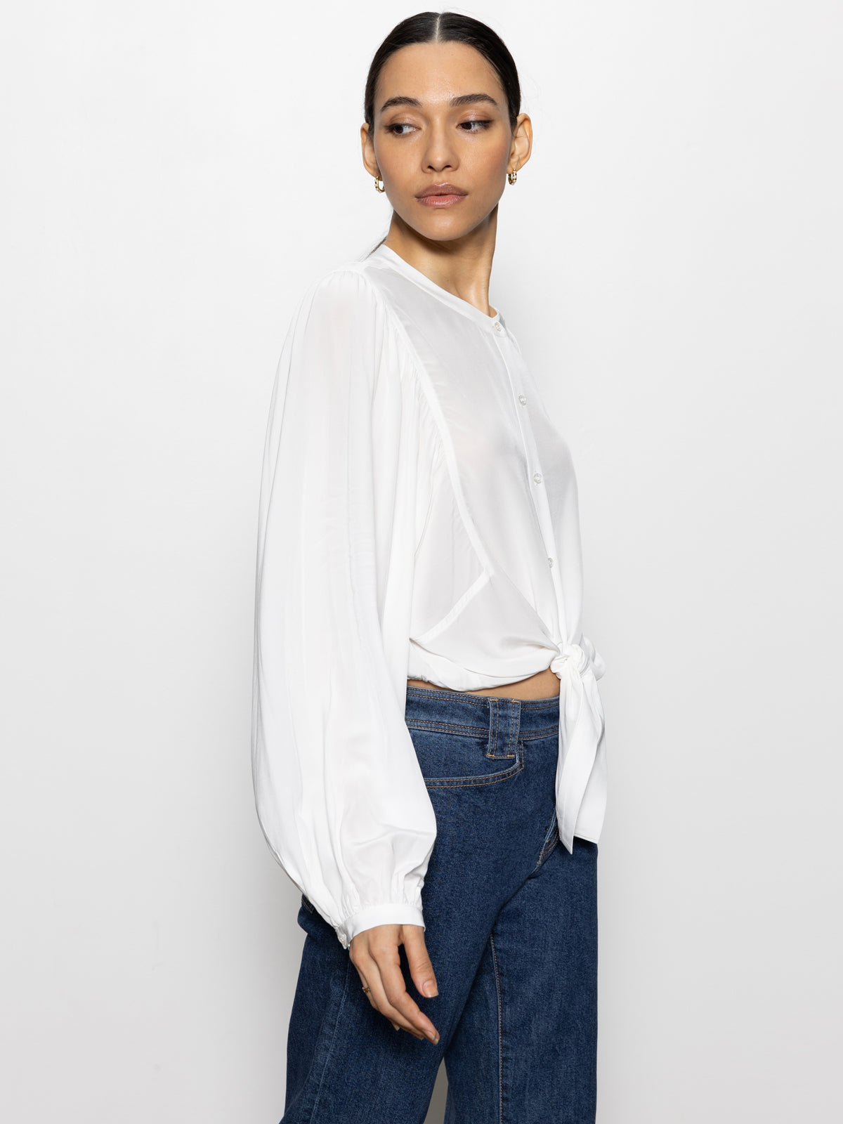 A person wearing the "Tie Front Volume Blouse Chalk" by Sanctuary Clothing, matched with blue jeans, is standing against a plain white background. The blouse features balloon sleeves and is tied at the waist. The individual has their hair pulled back and is looking slightly over their shoulder.