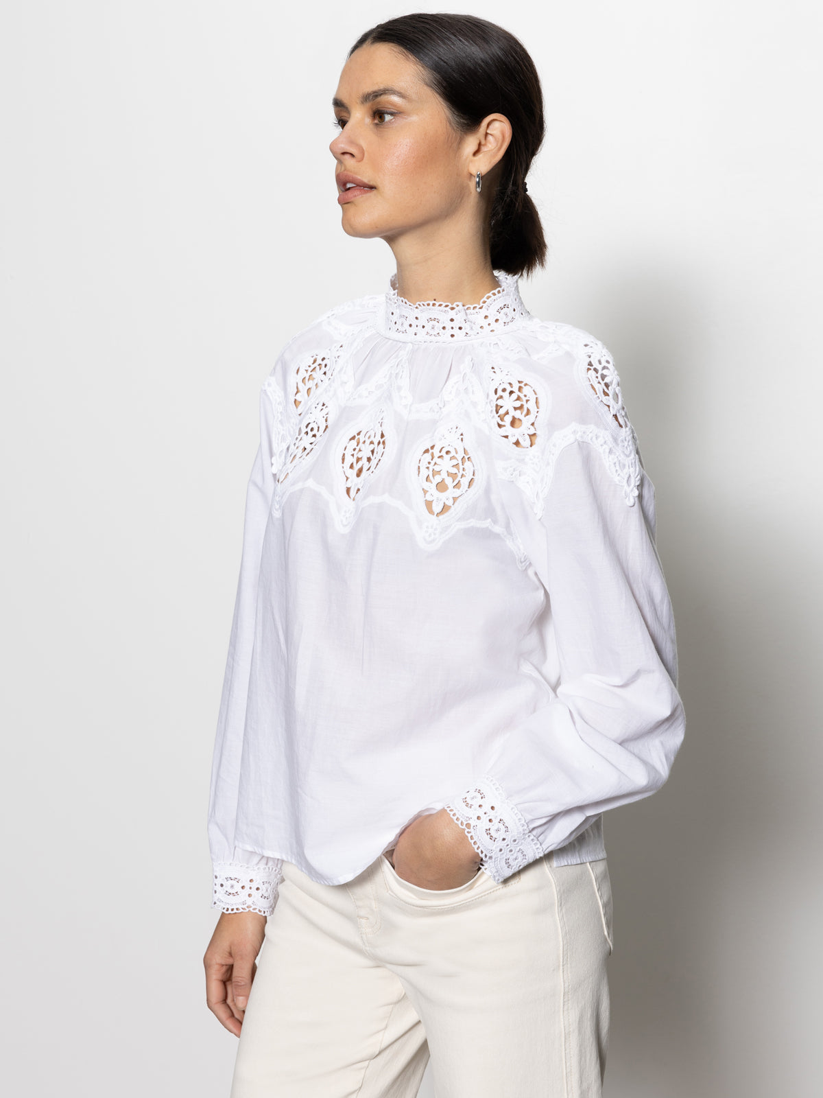 A person models the Sanctuary Clothing lace popover blouse in white against a plain backdrop. Their dark hair is pulled back as they look to the side, paired with light-colored pants.