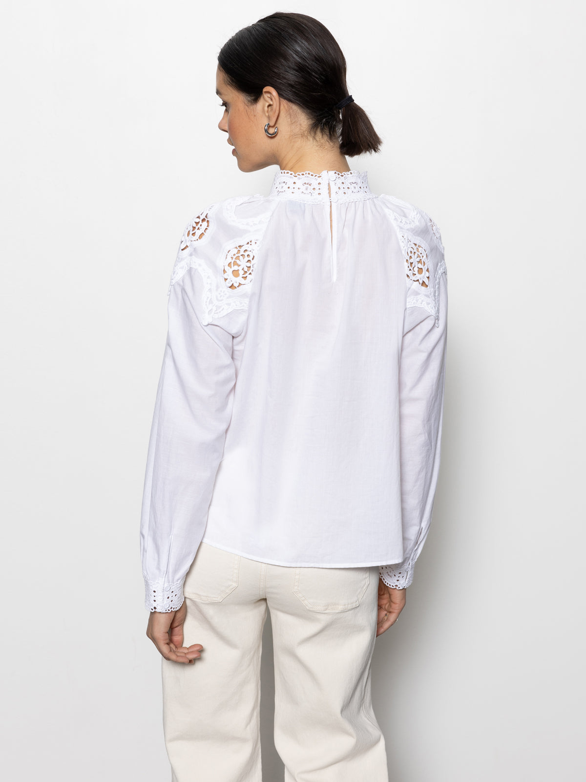 A dark-haired woman in a low bun is turned away, wearing Sanctuary Clothing's lace popover blouse in white. The blouse features intricate lace details on the shoulders and collar, with long sleeves gathered at the wrists. She complements it with cream-colored pants.