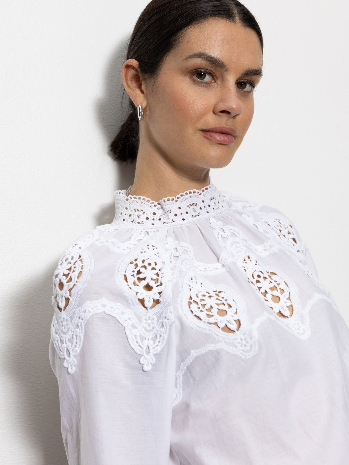 A person with dark hair is wearing the Sanctuary Clothing lace popover blouse in white, featuring intricate lace designs on the shoulders and collar. They're standing against a plain, light-colored background, looking slightly to the side.