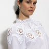 A person with dark hair is wearing the Sanctuary Clothing lace popover blouse in white, featuring intricate lace designs on the shoulders and collar. They're standing against a plain, light-colored background, looking slightly to the side.