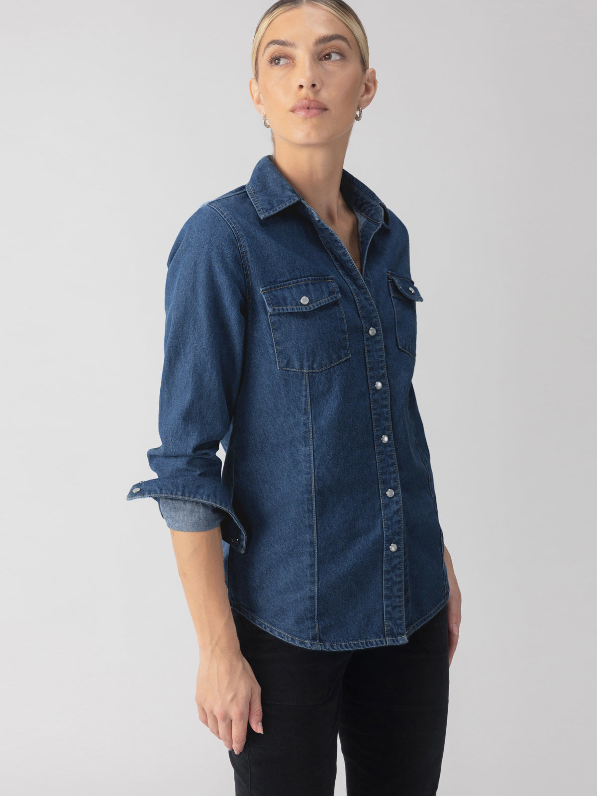 Person wearing The Denim Western Shirt Blue Slate by Sanctuary Clothing, with rolled-up sleeves and black pants, standing against a plain background, looking to the side.