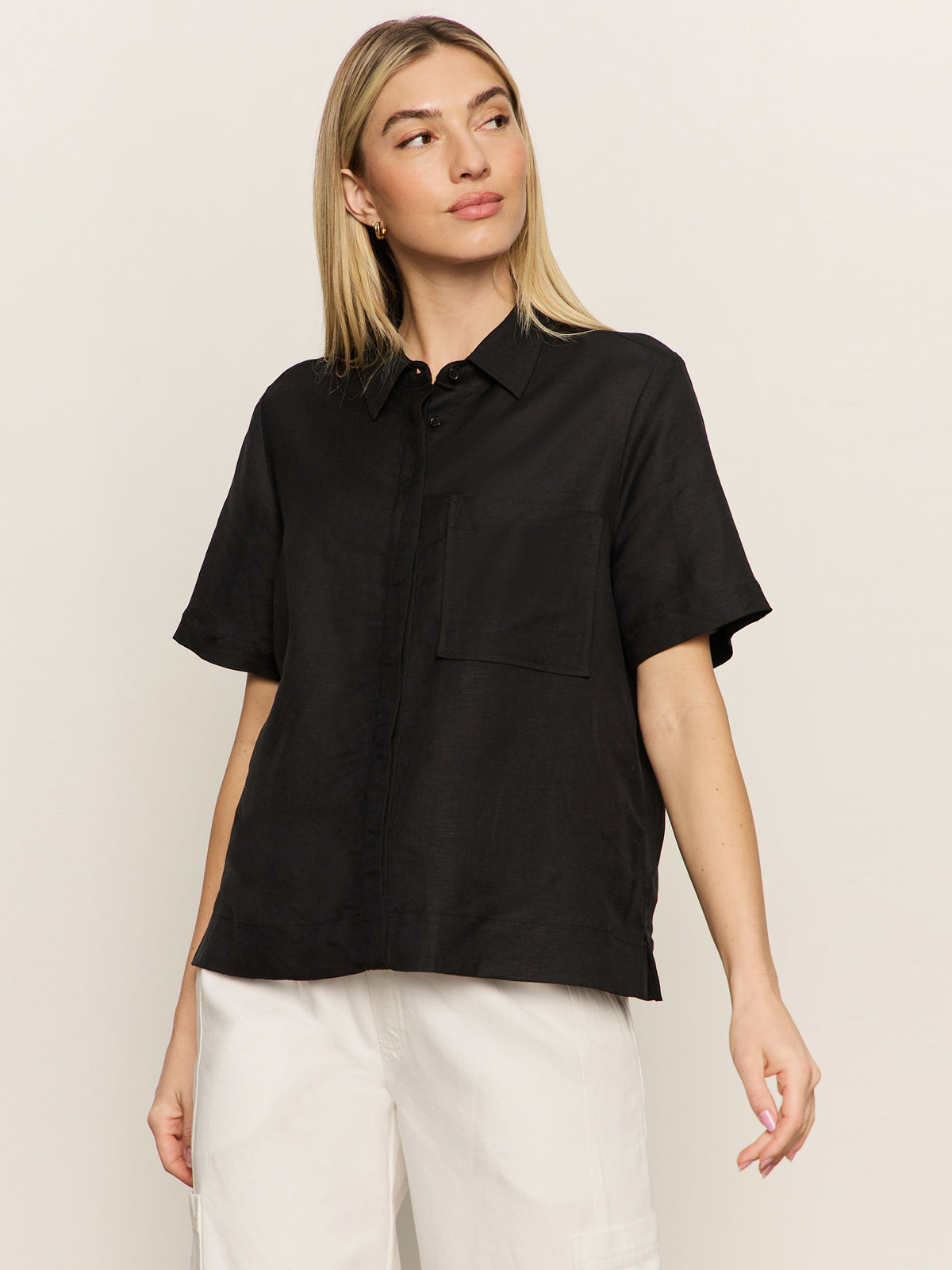 A woman with straight blonde hair wears the Sanctuary Clothing relaxed vacation shirt in black, a short-sleeve button-up with a chest pocket, paired with white pants, standing against a light background and looking slightly to the side.