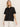 A woman with straight blonde hair wears the Sanctuary Clothing relaxed vacation shirt in black, a short-sleeve button-up with a chest pocket, paired with white pants, standing against a light background and looking slightly to the side.