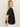 A woman with long blonde hair models the Sanctuary Clothing relaxed vacation shirt in black, paired with white pants, standing sideways and looking over her shoulder against a plain beige background.