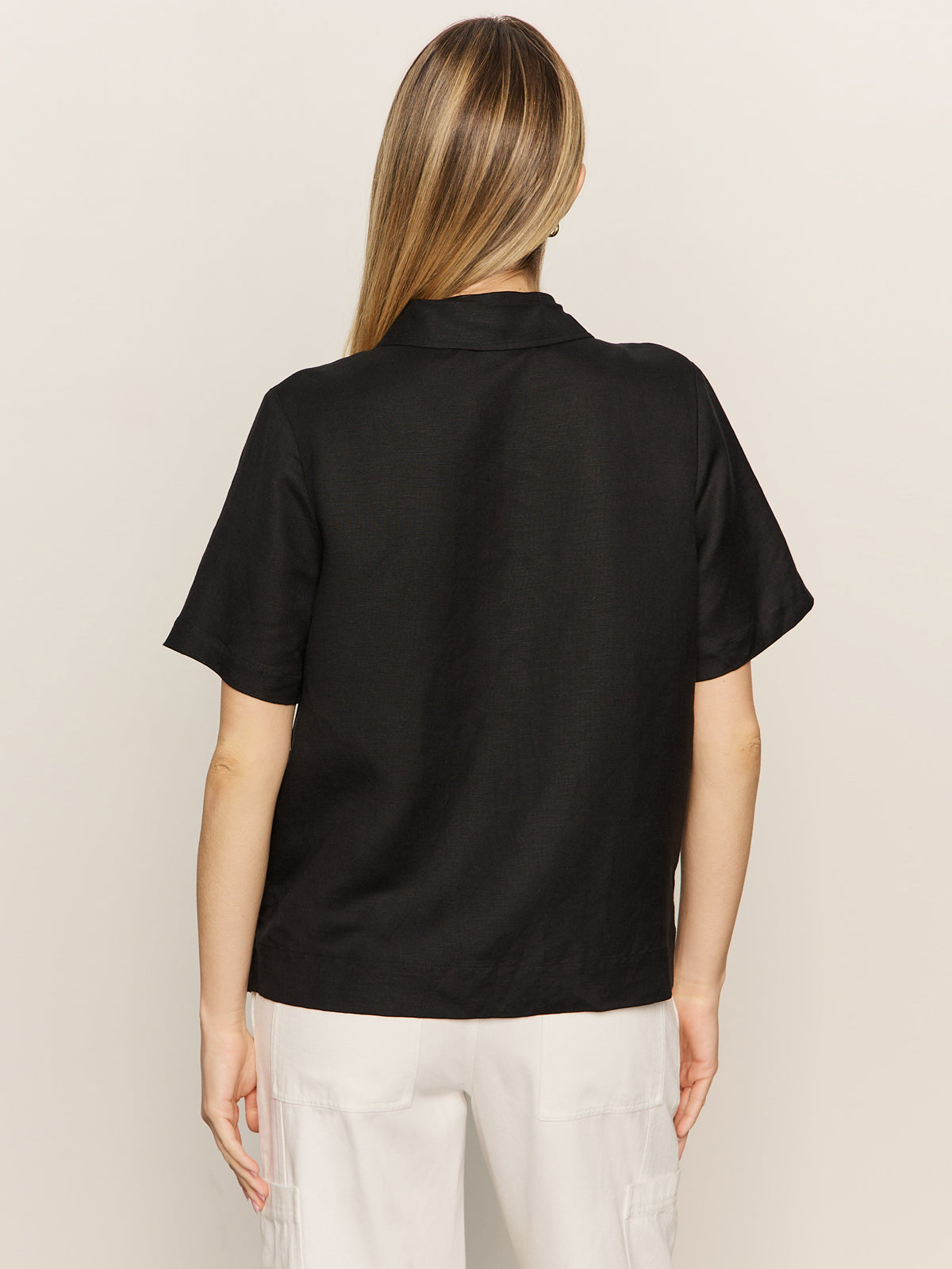 A woman with straight, light brown hair is shown from the back wearing the Sanctuary Clothing relaxed vacation shirt in black, paired with white pants against a plain light background.