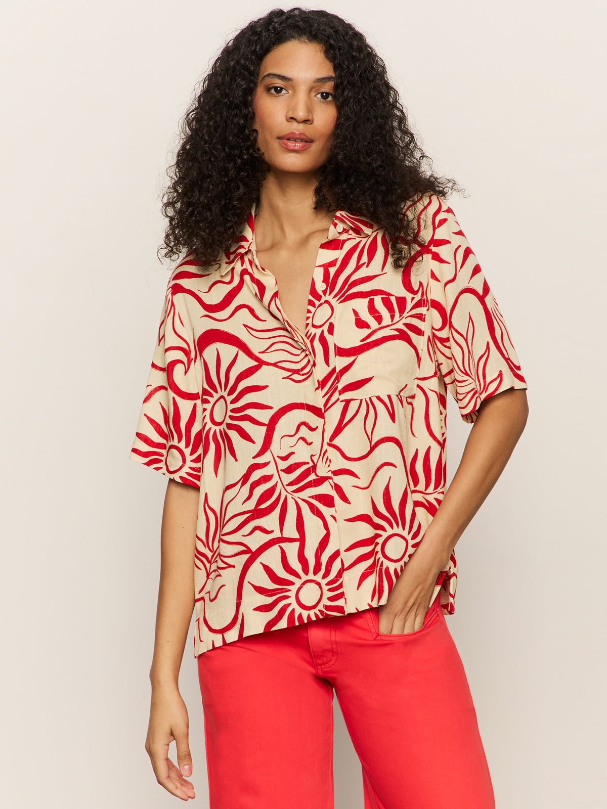 A woman with long curly hair wears the Sanctuary Clothing relaxed vacation shirt sunny riviera, featuring bold red sun and wave patterns on beige, paired with matching red pants against a plain light background.