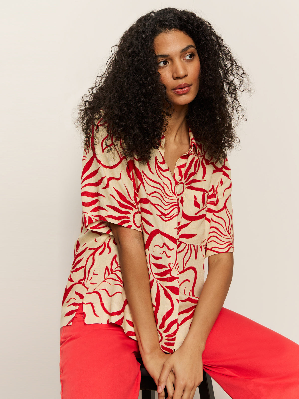 A woman with curly hair sits on a stool wearing the Sanctuary Clothing relaxed vacation shirt sunny riviera, featuring a beige base with a red abstract pattern, paired with bright red pants and looking slightly to the side against a plain background.