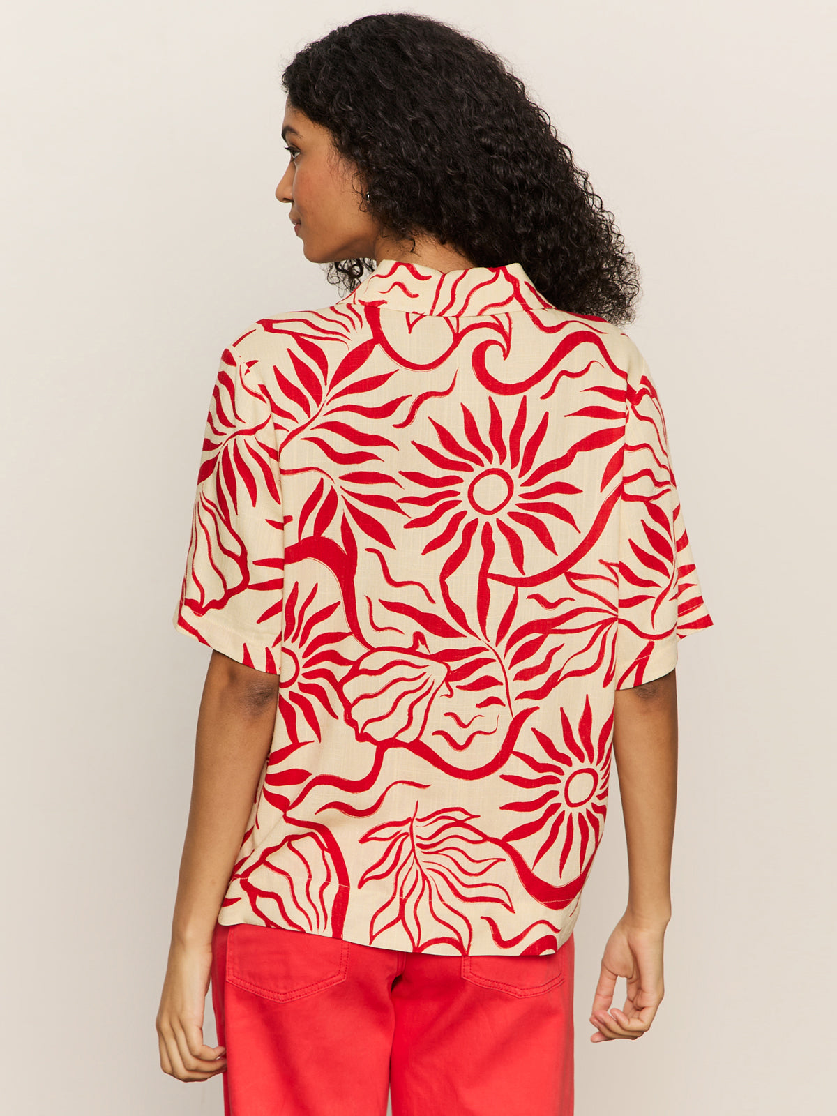 A person with curly hair wears the Sanctuary Clothing relaxed vacation shirt sunny riviera, featuring a bold red abstract sun and wave pattern on beige, paired with red pants. They face away from the camera against a plain background.