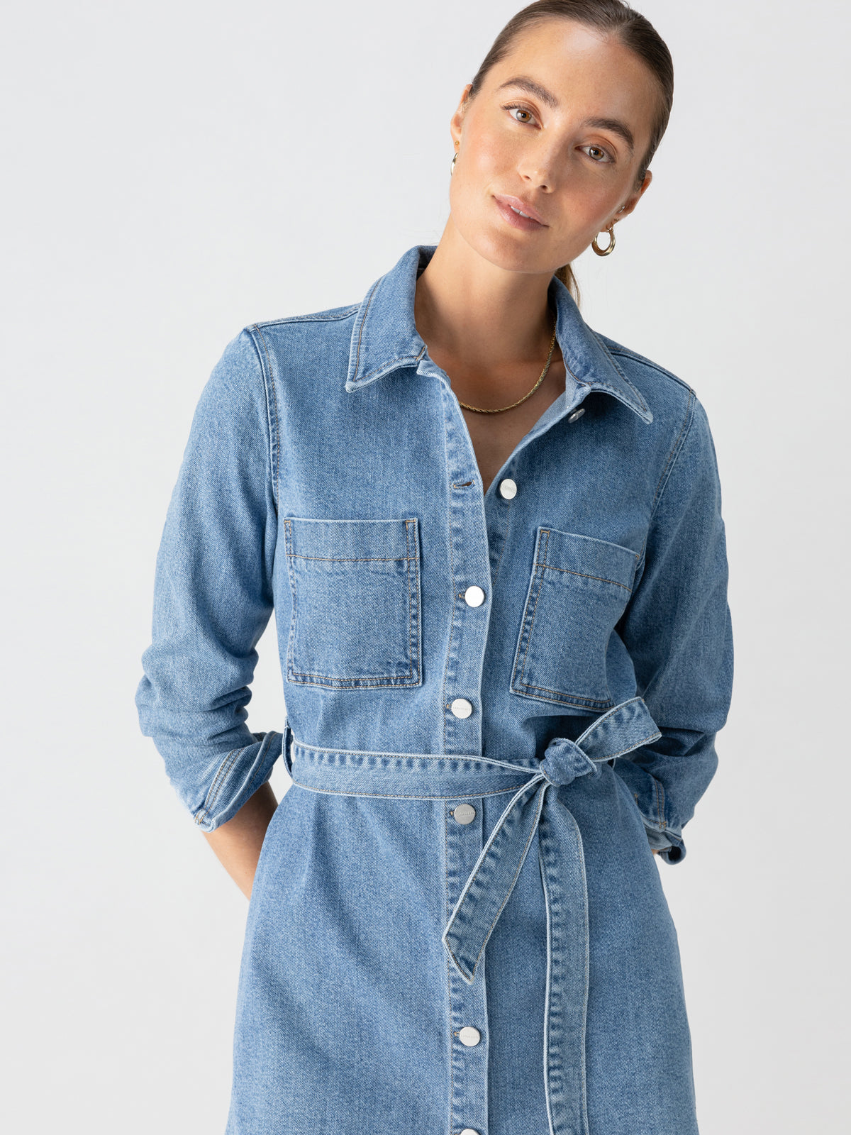 A person wearing the Denim Utility Shirt Dress Solar Flare by Sanctuary Clothing, featuring chest pockets and a tied belt, poses against a plain background. The individual has their hair pulled back, dons small hoop earrings, and maintains a calm expression.