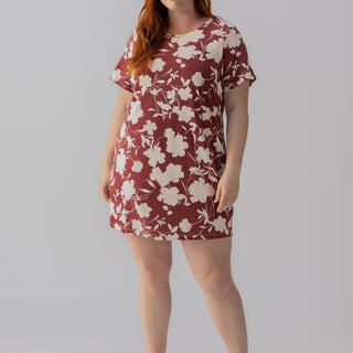 A woman with red hair is standing against a plain background. She is wearing "The Only One T Shirt Dress Warm Vista Inclusive Collection" by Sanctuary Clothing, which features a short, burgundy design with a large white floral pattern. She is also wearing tan sandals and smiling slightly.