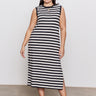 A person poses confidently in Sanctuary Clothing's contrast trim maxi dress in black macrame stripe, extended sizing, featuring side slits and paired with black sandals against a plain light-colored wall and floor.