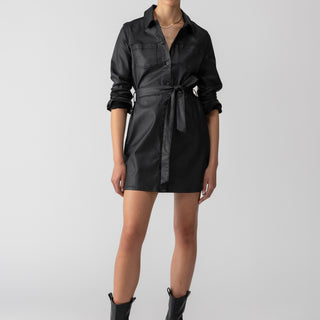 A person stands confidently wearing a Coated Denim Dress Black by Sanctuary Clothing, complete with a tied waist and black ankle boots. Their hair is pulled back, and they accessorize with small earrings and a delicate necklace. The background is a plain light gray.