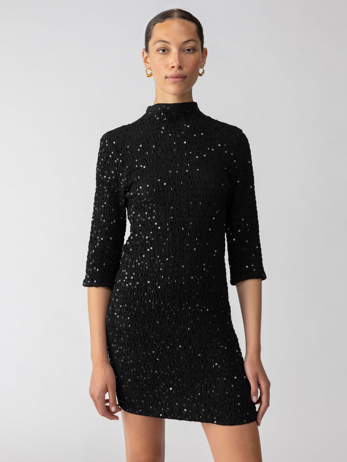 A person with their hair pulled back is wearing the Holiday Dream Smocked Sequin Dress in Black by Sanctuary Clothing, a stunning mini dress featuring three-quarter sleeves and a high neckline. They accessorize with gold hoop earrings and stand against a plain grey background.