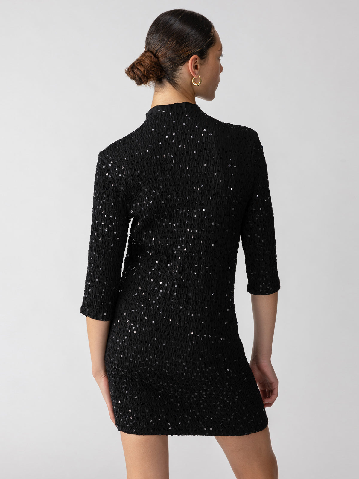 A woman with brown hair in a bun is wearing the Holiday Dream Smocked Sequin Dress in Black by Sanctuary Clothing, featuring a sparkly, black, long-sleeve design with a high neckline. She is facing away from the camera, highlighting the back of the dress against a plain, light-colored background.