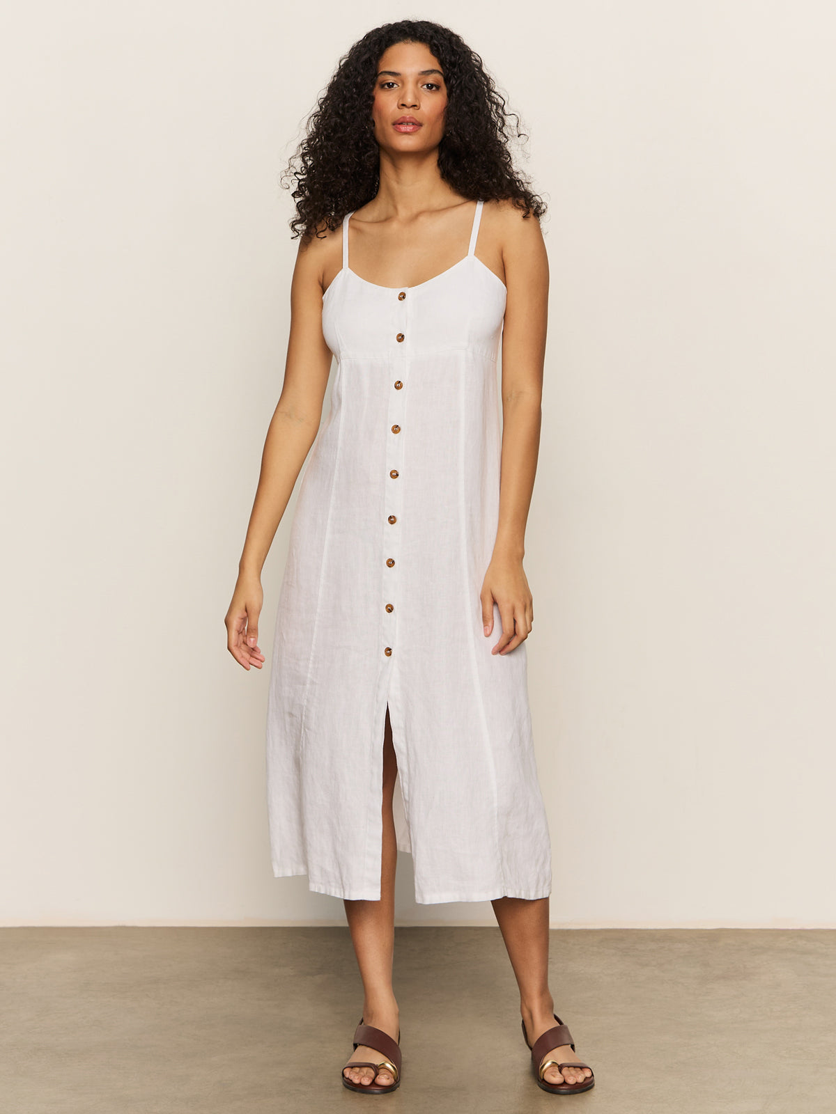 A woman with curly hair wears a Sanctuary Clothing Wilder Linen Dress in bleached white, featuring a sleeveless button-down design with spaghetti straps, standing against a plain background. She completes the look with brown sandals.