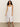 A woman with curly hair wears a Sanctuary Clothing Wilder Linen Dress in bleached white, featuring a sleeveless button-down design with spaghetti straps, standing against a plain background. She completes the look with brown sandals.